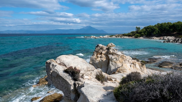 Wybrzeże Morza Egejskiego otoczone zielenią, skały, krzewy i drzewa, błękitna woda z falami, góra