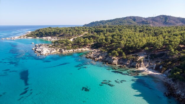 Wybrzeże Morza Egejskiego z niebieską przezroczystą wodą, zielenią dookoła, skałami, krzewami i drzewami, widok z drona, Grecja