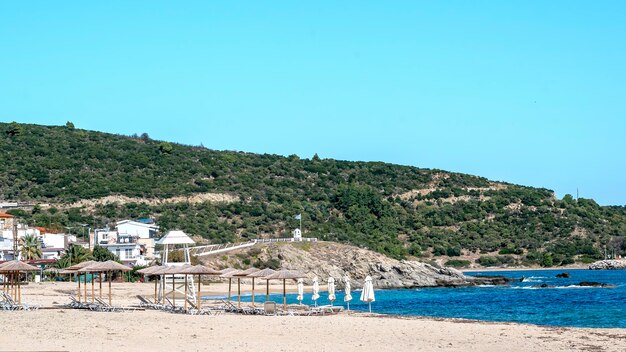 Wybrzeże Morza Egejskiego z zabudowaniami po lewej stronie, skały, parasole z leżakami, krzewy i drzewa, błękitna woda ze wzgórzem w Sarti w Grecji