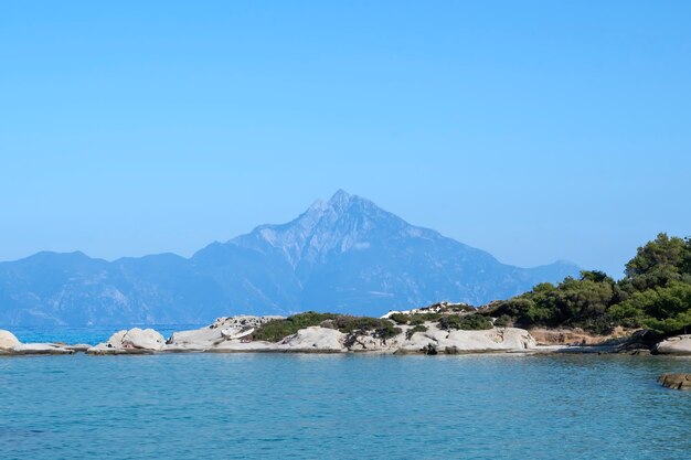 Wybrzeże Morza Egejskiego ze skałami i odpoczywającymi ludźmi, góry w oddali, zieleń na pierwszym planie, błękitna woda, Grecja