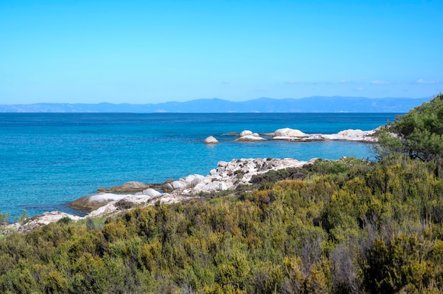 Wybrzeże Morza Egejskiego ze skałami nad wodą i lądem w oddali, zieleń na pierwszym planie, błękitna woda, Grecja
