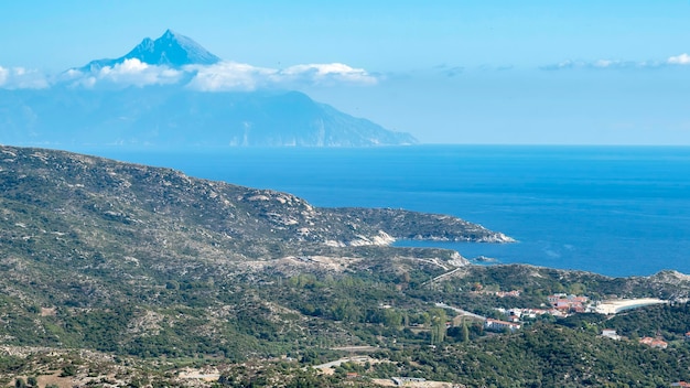 Wybrzeże Morza Egejskiego ze wzgórzami pełnymi zieleni, zabudowania w pobliżu wybrzeża z wysoką górą sięgającą chmur Grecja
