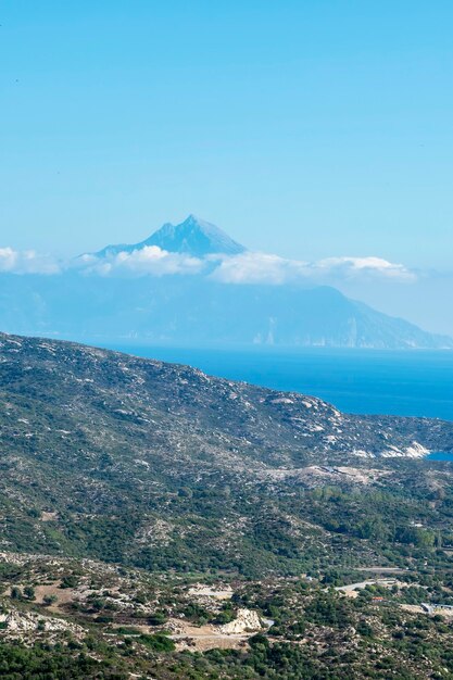 Wybrzeże Morza Egejskiego ze wzgórzami pełnymi zieleni, zabudowania w pobliżu wybrzeża z wysoką górą sięgającą chmur Grecja