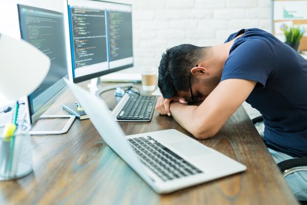Wyczerpany młody mężczyzna programista śpiący przy technologii, opierając się o biurko