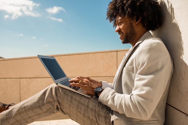 Wygodny Afroamerykanin pracujący na świeżym powietrzu. Mężczyzna w garniturze z brodą za pomocą laptopa. Siedząc na tarasie lub na dachach. Praca, menedżer, koncepcja technologii