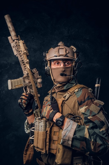 Wykonawca prywatnej ochrony w mundurze kamuflażu pozuje z karabinem szturmowym. Zdjęcie studyjne na tle ciemnej, teksturowanej ściany