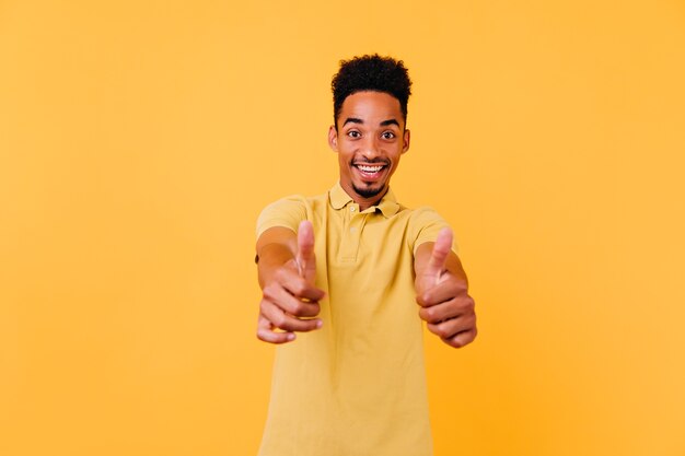 Wyrafinowany czarny chłopiec pokazujący kciuki do góry z zaskoczonym uśmiechem. Kryty zdjęcie wesołego afrykańskiego mężczyzny z zabawną fryzurą.