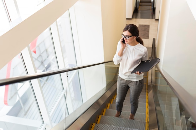 Wysoki kąt bizneswoman z segregatorem rozmawia przez telefon na schodach ruchomych