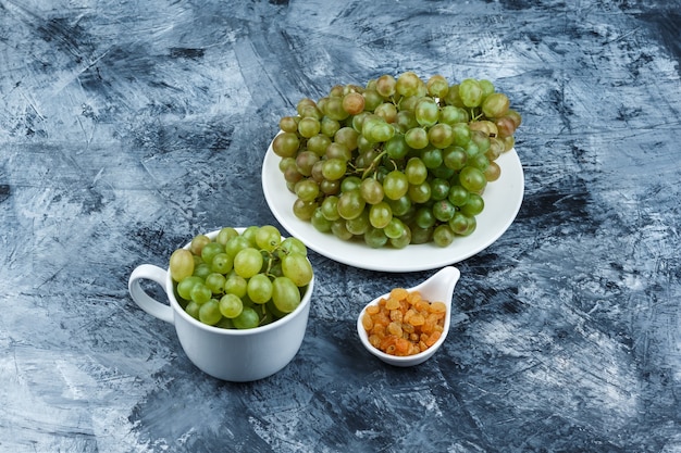 Bezpłatne zdjęcie wysoki kąt widzenia zielone winogrona w białej płytce i kubek z rodzynkami na tle nieczysty tynku. poziomy