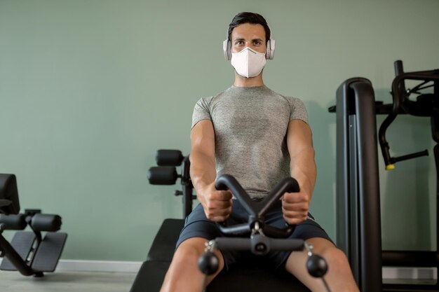 Wysportowany mężczyzna z maską na twarz, który trenuje na maszynie do wiosłowania na siłowni