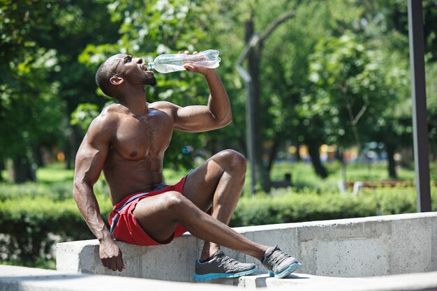 Wysportowany sportowiec odpoczywa i pije wodę po ćwiczeniach na stadionie. Afro lub Afroamerykanin mężczyzna na zewnątrz w mieście. fitness, zdrowie, koncepcja stylu życia