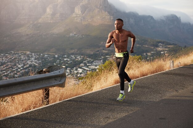 Wysportowany zdrowy człowiek biegnie wzdłuż drogi na zewnątrz, pokonuje duże odległości, przygotowuje się do maratonu. Sportowy mężczyzna ćwiczy z górki, nosi sportowe buty, legginsy, jest w dobrej formie
