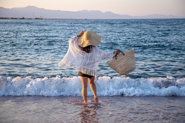 Wzdłuż wybrzeża spaceruje dziewczyna w wielkim kapeluszu i wiklinowej torbie. koncepcja wakacji letnich.