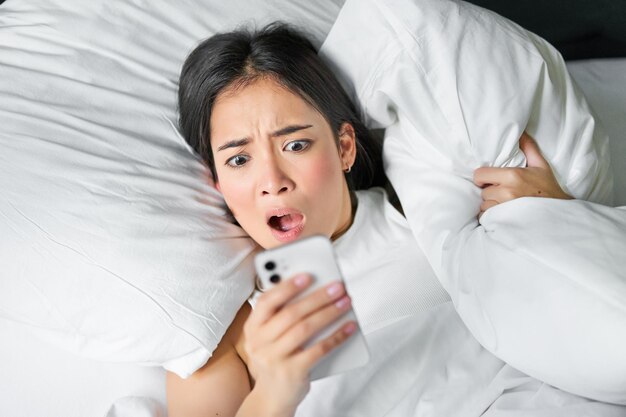 Z bliska portret azjatyckiej dziewczyny leżącej w łóżku, patrzącej na smartfona, który budzi się późno i gwiazda