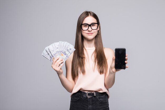 Z podnieceniem młoda amerykańska kobieta trzyma fan dolarowi rachunki i srebny telefon komórkowy odizolowywający nad biel ścianą