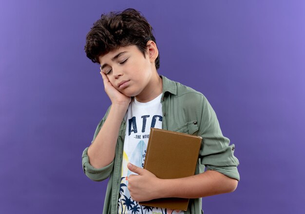 Z zamkniętymi oczami chory mały uczeń trzyma książkę i kładzie rękę na policzku na białym tle na fioletowej ścianie