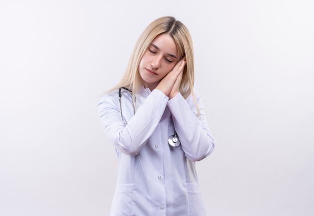 Z zamkniętymi oczami lekarz młoda blondynka ubrana w stetoskop i fartuch medyczny pokazujący gest snu na odosobnionej białej ścianie