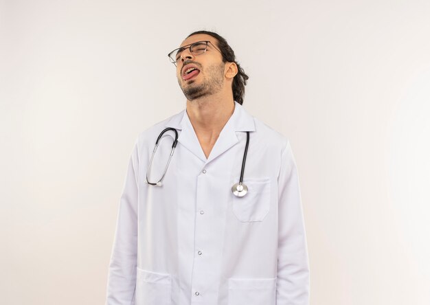 Z zamkniętymi oczami młody lekarz płci męskiej z okularami optycznymi, ubrany w białą szatę ze stetoskopem, pokazujący język na odosobnionej białej ścianie z miejscem na kopię