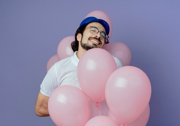 Z zamkniętymi oczami radosny przystojny mężczyzna w okularach i niebieskim kapeluszu trzymając balony na fioletowym tle