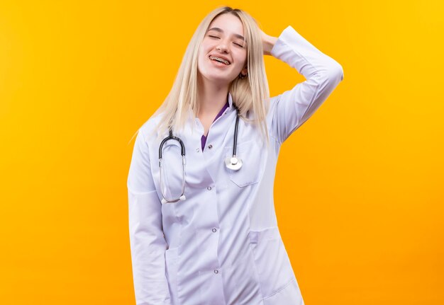 Z zamkniętymi oczami uśmiechnięta lekarz młoda dziewczyna ubrana w stetoskop w fartuchu medycznym i aparat ortodontyczny położyła dłoń na głowie na odizolowanej żółtej ścianie