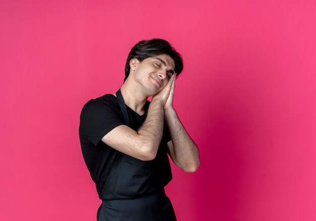 Z zamkniętymi oczami zadowolony młody przystojny mężczyzna fryzjer w mundurze pokazujący gest snu na różowym tle