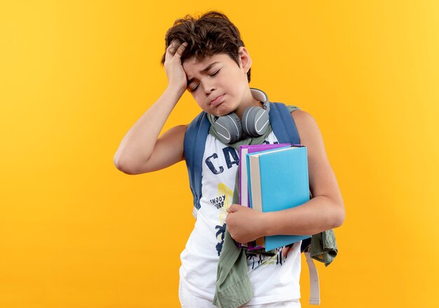 Z zamkniętymi oczami zmęczony mały chłopiec w wieku szkolnym, ubrany w plecak i słuchawki, trzymający książki i chwycił głowę odizolowaną na żółto