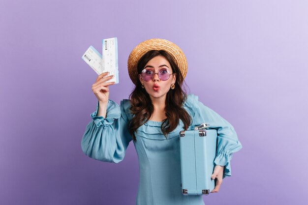 Zabawna kobieta w marynarce i liliowych okularach patrzy ze zdumieniem, pokazując swoje bilety lotnicze i niebieską walizkę retro.
