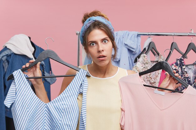 Zabawna, zszokowana młoda kobieta z opaską trzymającą w obu rękach dwa wieszaki z modnymi ubraniami, która ma ochotę kupić je obie podczas zakupów w sklepie na wielkiej wyprzedaży. Koncepcja konsumpcjonizmu