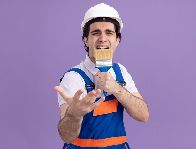 Zabawny i radosny młody konstruktor w mundurze konstrukcyjnym i kasku ochronnym, trzymając pędzel, używając jako śpiew mikrofonu stojącego nad fioletową ścianą