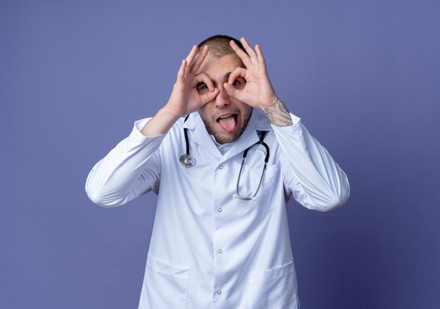 Zabawny młody lekarz płci męskiej ubrany w szlafrok medyczny i stetoskop na szyi, wykonujący gest spojrzenia, używając rąk jako lornetki i pokazując język odizolowany na fioletowym tle z przestrzenią do kopiowania