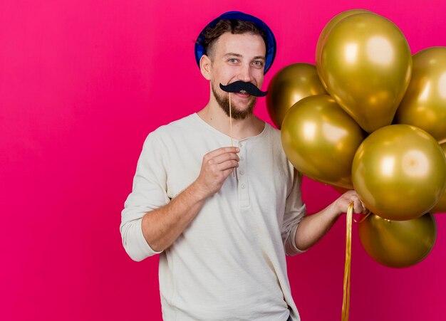Zabawny uśmiechnięty młody przystojny słowiański facet w kapeluszu imprezowym, trzymając balony i fałszywe wąsy na patyku nad ustami, patrząc z przodu na białym tle na różowej ścianie z miejscem na kopię
