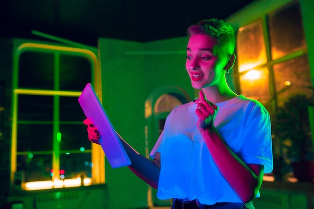 Bezpłatne zdjęcie zachwycający. kinowy portret stylowej kobiety w oświetlonym neonem wnętrzu. stonowane jak efekty kinowe, jasne neonowane kolory. model kaukaski za pomocą tabletu w kolorowych światłach w pomieszczeniu. kultura młodzieżowa.