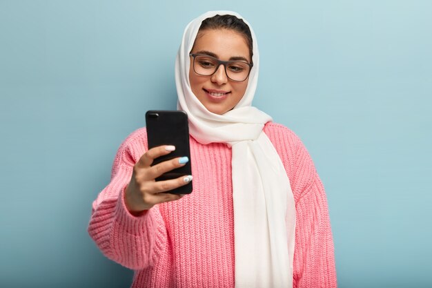 Zachwycona muzułmanka z delikatnym uśmiechem, trzyma telefon z przodu, robi selfie, nosi prostokątne okulary i welon, ma manicure. Czas na zrobienie zdjęcia