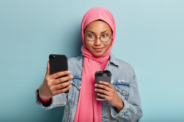 Zachwycona, Przyjemnie Wyglądająca Muzułmańska Dziewczyna Pije Kawę Na Wynos, Robi Selfie Lub Prowadzi Rozmowę Wideo, Ubrana W Stylową Dżinsową Kurtkę I Hidżab, Udostępnia Zdjęcia Obserwującym W Sieciach Społecznościowych