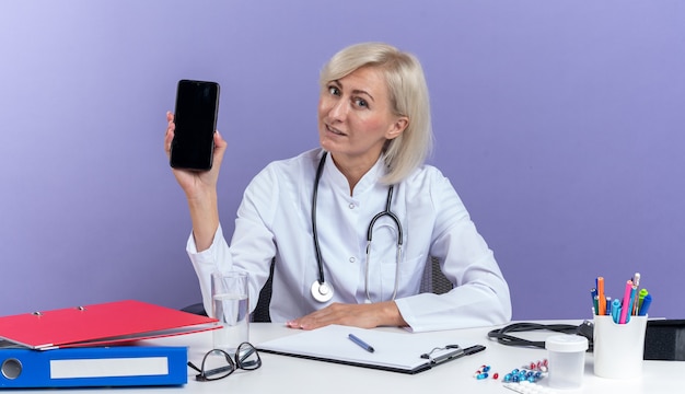 Zadowolona dorosła słowiańska lekarka w szacie medycznej ze stetoskopem siedzi przy biurku z narzędziami biurowymi trzymając telefon na białym tle na fioletowym tle z kopią przestrzeni