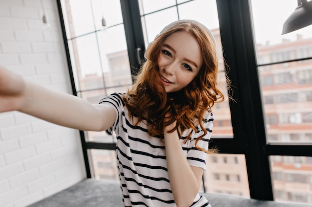 Zadowolona dziewczyna z rudymi kręconymi włosami robi sobie zdjęcie. Kryty strzał z ładną kobietą Dokonywanie selfie.
