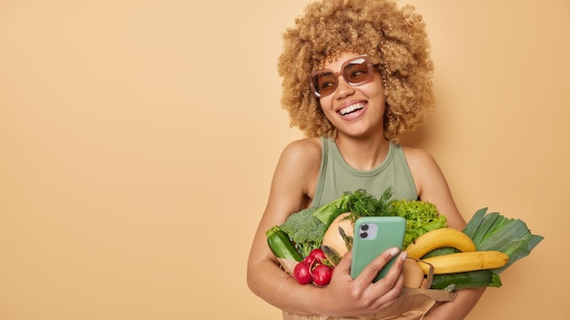 Bezpłatne zdjęcie zadowolona kobieta używa telefonu komórkowego do zamawiania produktów online, nosi świeże warzywa i owoce kupione na targu, nosi okulary przeciwsłoneczne i koszulkę odizolowane na brązowym tle, puste miejsce na promocję