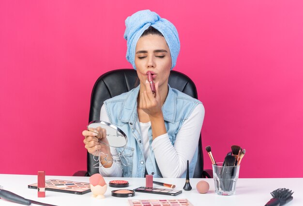 Zadowolona ładna kaukaska kobieta z zawiniętymi włosami w ręcznik, siedząca przy stole z narzędziami do makijażu, trzymająca lustro i nakładająca szminkę na różowej ścianie z kopią przestrzeni