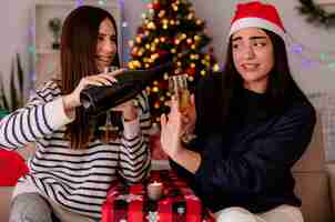 Bezpłatne zdjęcie zadowolona ładna młoda dziewczyna w czapce świętego mikołaja trzyma kieliszek szampana i gesty znak stop dla swojej przyjaciółki trzymającej butelkę szampana siedzącej na fotelu i cieszącej się świątecznymi chwilami w domu