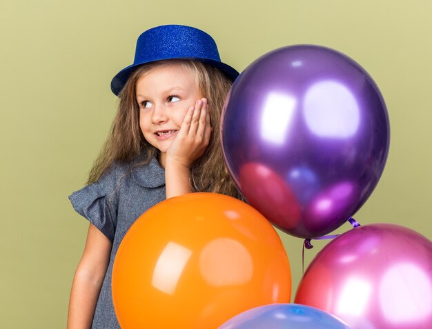 zadowolona mała blondynka w niebieskim kapeluszu imprezowym stojąca z balonami z helem kładąca dłoń na twarzy i patrząca w bok odizolowana na oliwkowozielonej ścianie z kopią przestrzeni