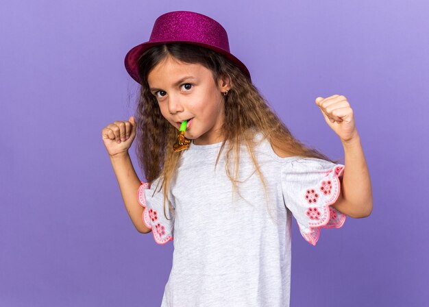 zadowolona mała kaukaska dziewczyna w fioletowym kapeluszu imprezowym dmuchająca w gwizdek i trzymająca pięści odizolowane na fioletowej ścianie z miejscem na kopię