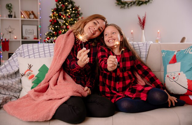 zadowolona mama i córka trzymają zimne ognie przykryte kocem, siedząc na kanapie i ciesząc się świętami Bożego Narodzenia w domu