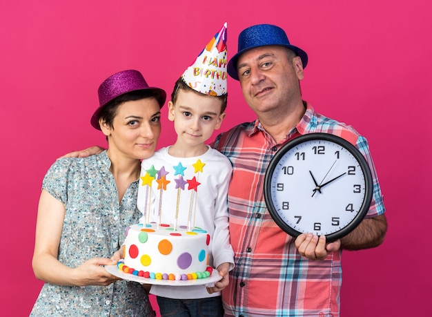 Zadowolona Matka W Fioletowej Czapce Trzymającej Tort Urodzinowy I Uśmiechnięty Ojciec W Niebieskiej Imprezowej Czapce Trzymający Zegar Stojący Z Synem Odizolowany Na Różowej ścianie Z Kopią Miejsca