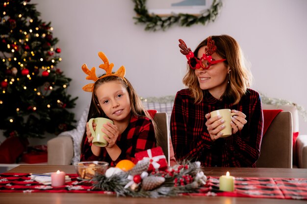 zadowolona matka w reniferowych okularach patrząca na córkę trzymającą filiżankę siedzącą przy stole cieszącą się świętami Bożego Narodzenia w domu