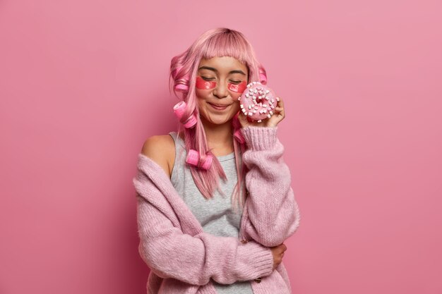 Zadowolona młoda Azjatka o różowych włosach, zamyka oczy, trzyma pyszne pączki przy twarzy, nakłada kolagenowe łaty pod oczy, tworzy kręconą fryzurę