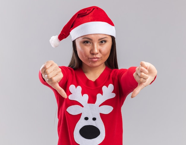 Zadowolona młoda azjatycka dziewczyna ubrana w świąteczny kapelusz ze swetrem pokazując kciuki w dół na białym tle