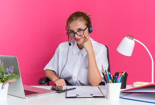 Zadowolona młoda blondynka call center dziewczyna nosi zestaw słuchawkowy i okulary, siedząc przy biurku z narzędziami do pracy, wskazując palcem na policzek, patrząc na kamerę odizolowaną na różowej ścianie