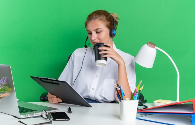 Zadowolona młoda blondynka call center dziewczyna nosi zestaw słuchawkowy, siedząc przy biurku z narzędziami do pracy, trzymając plastikową filiżankę kawy i schowek, patrząc na schowek na białym tle na zielonej ścianie