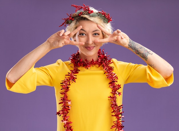 Bezpłatne zdjęcie zadowolona młoda blondynka ubrana w świąteczny wieniec na głowę i świecącą girlandę na szyi, patrząc na kamerę pokazującą symbole v w pobliżu oczu odizolowanych na fioletowym tle