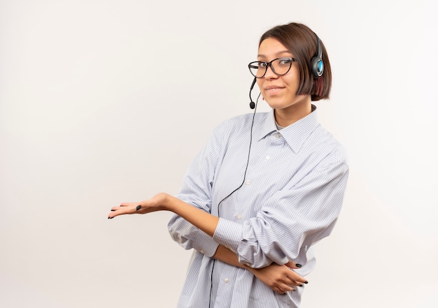 Zadowolona młoda dziewczyna call center w okularach i zestaw słuchawkowy pokazując pustą rękę na białym tle na białym tle z miejsca na kopię